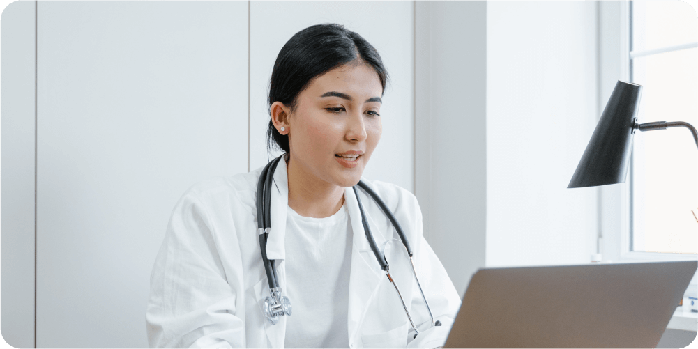 Женщина-врач в белом халате со стетоскопом на шее сидит за столом. Она сосредоточенно смотрит на экран ноутбука, демонстрируя профессионализм и сосредоточенность.