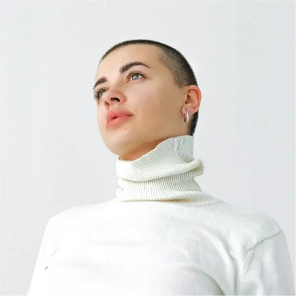 Человек с бритой головой и серьгами-кольцами смотрит вверх с безмятежным выражением лица. На нём белая водолазка на однотонном фоне.