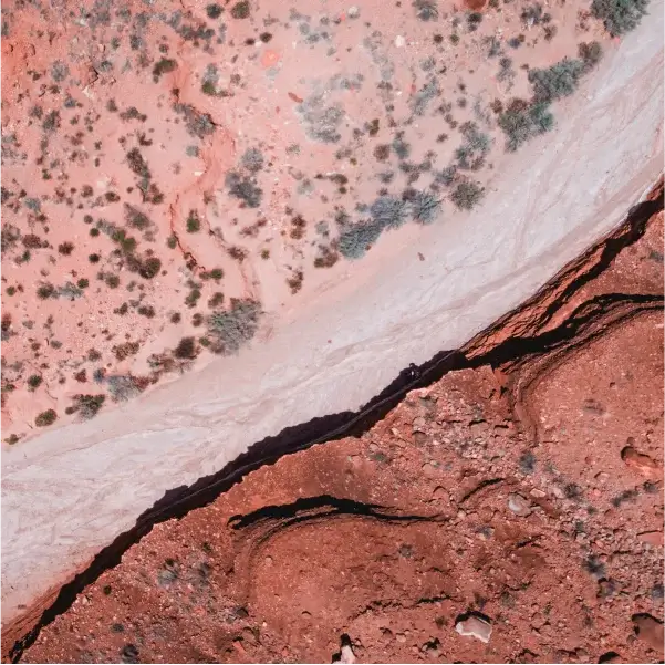 Вид с воздуха на каменистый пустынный ландшафт с красно-белыми полосами. Редкая растительность усеивает местность. Сцена создаёт ощущение засушливого одиночества.