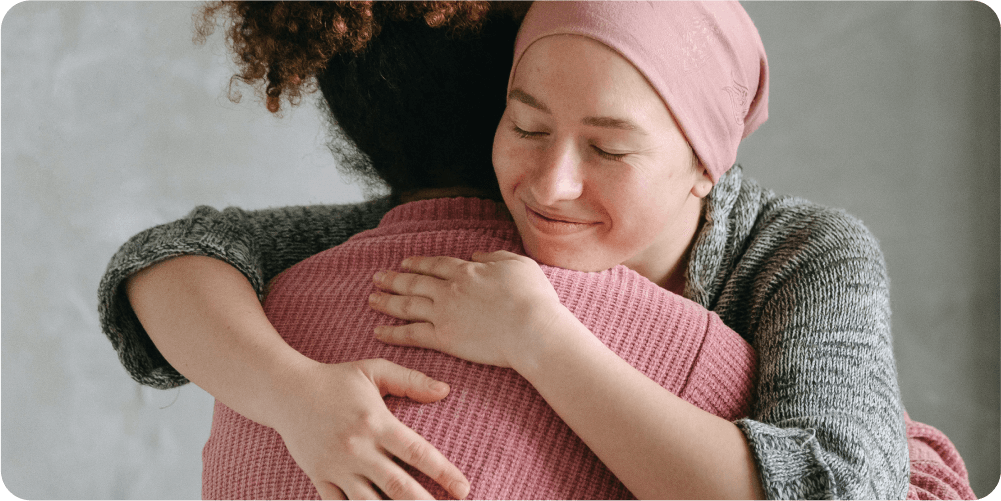 Две женщины тепло обнимаются, одна из них в платке, выражая утешение и поддержку. Спокойная обстановка, мягкое освещение и нейтральные тона.