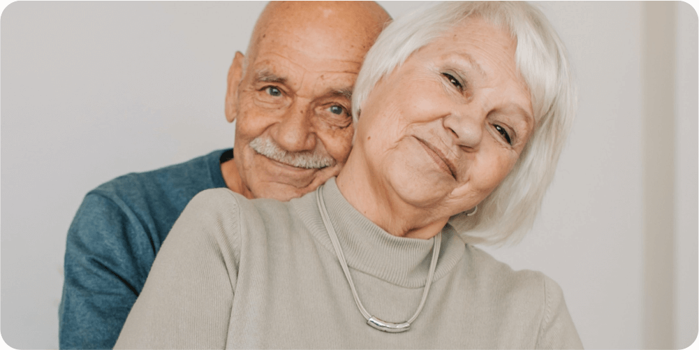 Пожилая пара тепло улыбается, мужчина нежно обнимает женщину сзади. Они излучают счастье и нежность на фоне однотонной фотографии.