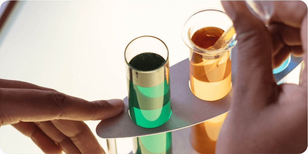 Руки держат набор пробирок с зелёными и оранжевыми жидкостями на белом фоне. Сцена создаёт научный, исследовательский тон.