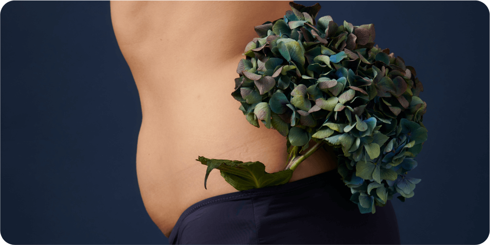 Крупный план торса человека на тёмно-синем фоне. На нём тёмно-синие шорты, за пояс заткнут букет синих гортензий.