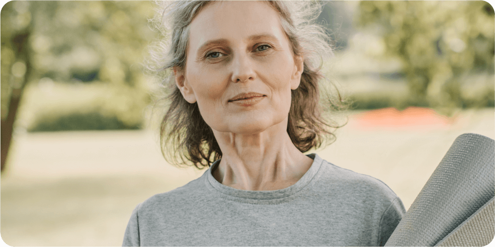 Пожилая женщина с короткими седыми волосами стоит снаружи, держа коврик для йоги, в серой футболке. Она выглядит уверенной и спокойной на фоне зелени.